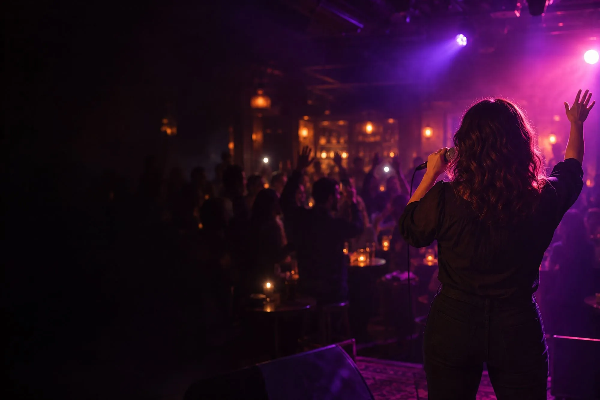 A singer seen from behind on a small bar stage, one arm raised to a cheering crowd under violet and pink lights
