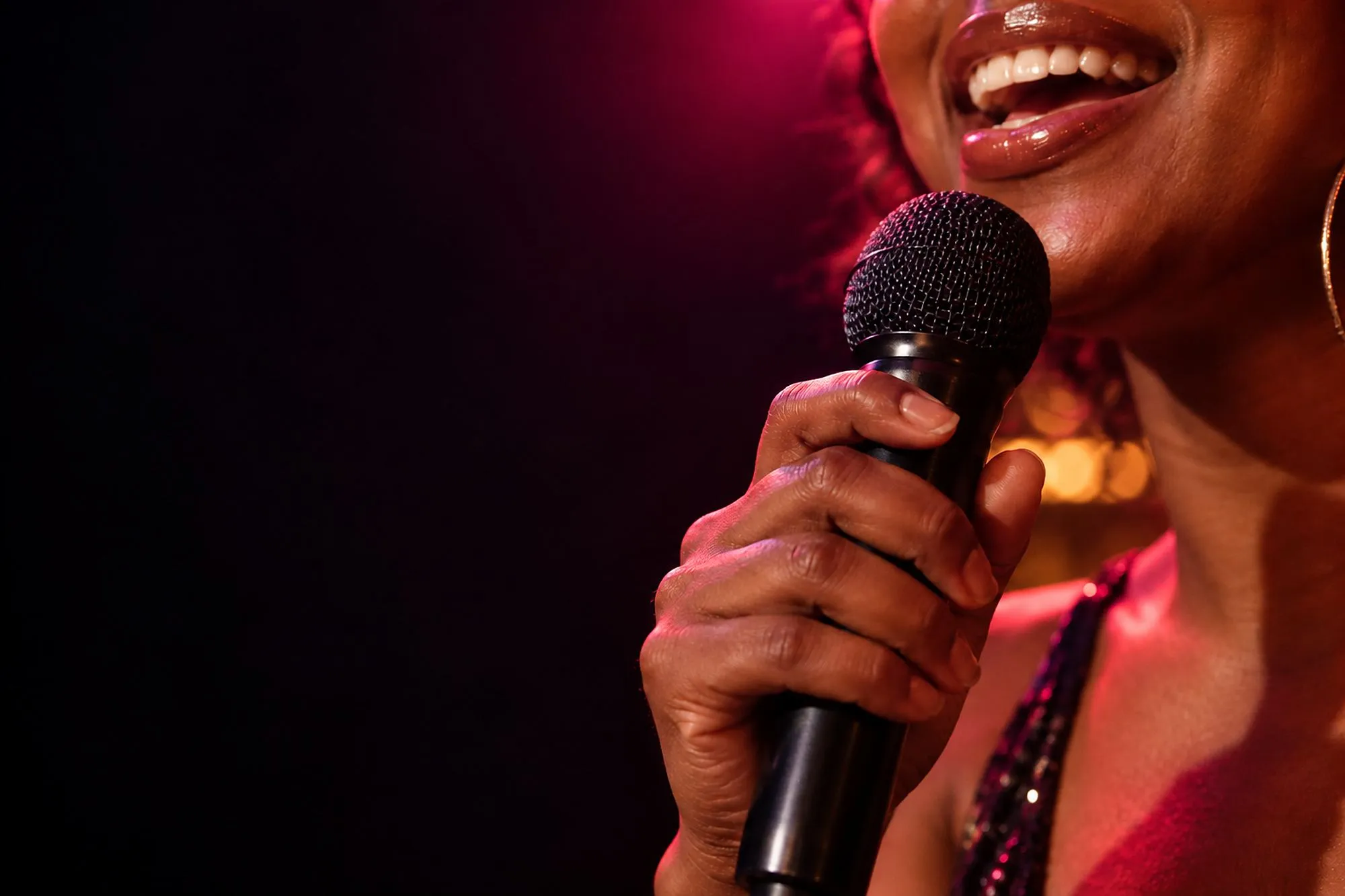 A smiling singer’s hand grips a microphone close to their mouth in warm pink light