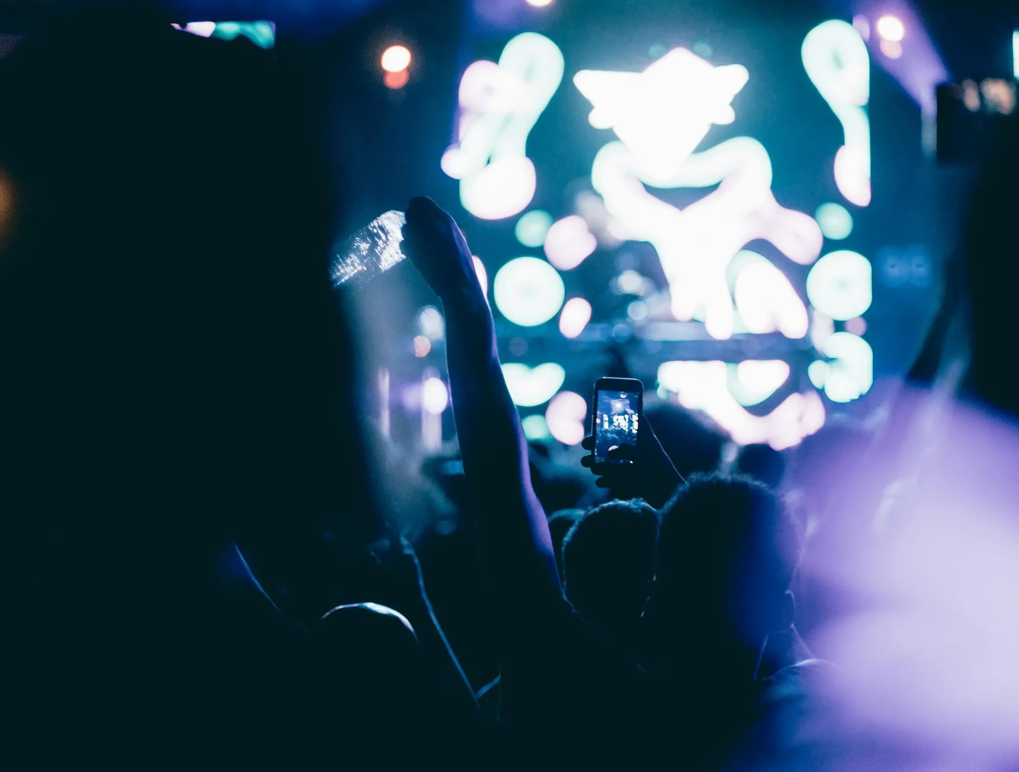 An audience seen from behind, an arm and a phone raised toward a glowing stage screen